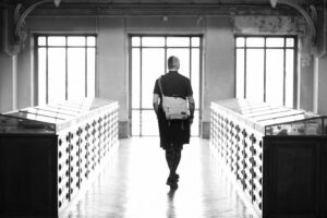 A black and white photo of Rob (aka mayhem) walking away from the camera and into the light, in an old building. Even from behind you can see his unforgettable hair and dress style!